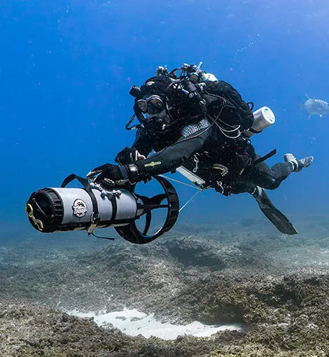 Two divers on closed-circuit rebreathers riding diver propulsion vehicles above a reef